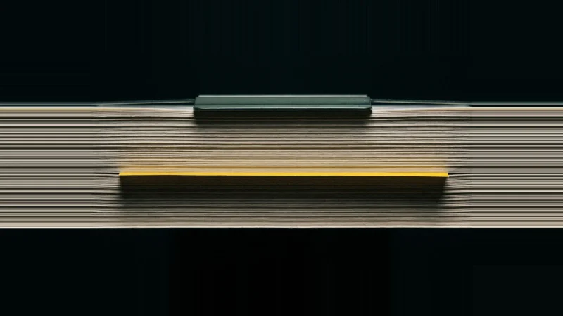 Macro view of a paper stack seen edge-on, one sheet pushed proud with its edge lit yellow against black.