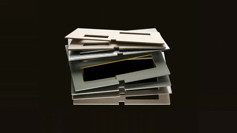 Overhead exploded stack of drawing-sheet plates in paper, concrete and metal, one slot empty and edged in yellow.