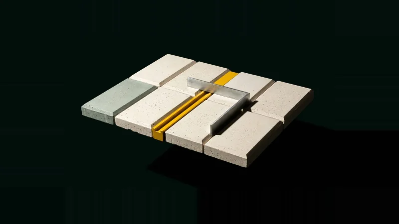 Overhead view of fitted pale concrete tiles with one narrow gap holding a yellow machined bar, on black.