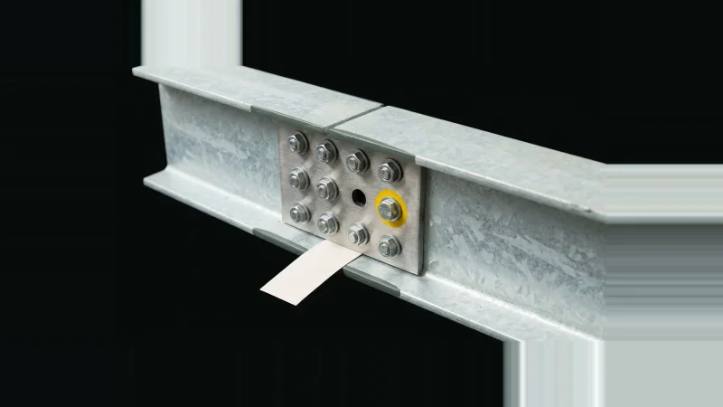 Close low-angle view of a bolted steel beam splice plate with one empty bolt hole and a yellow-marked washer on black.