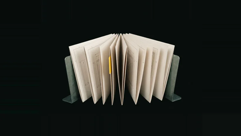 A fanned row of pale document packages on edge, one slot empty, marked by a single yellow tab in darkness.