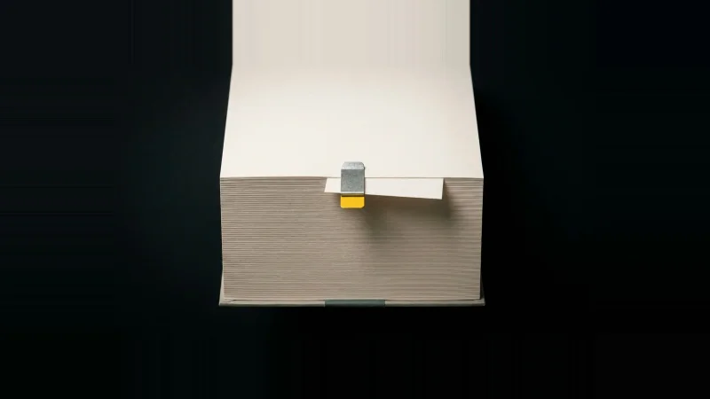 Overhead view of a thick off-white spec paper stack with one slip wedged out of alignment, tagged yellow.