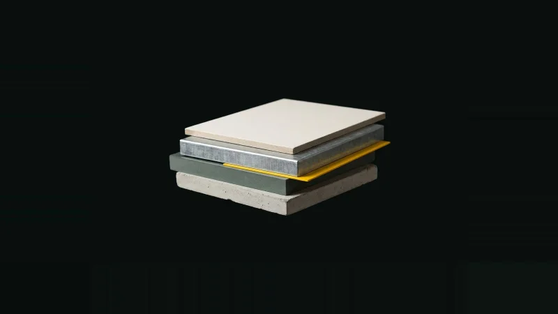 Low-angle photo of four small offset slabs of concrete, grey-green, metal and paper stacked in darkness, one seam shimmed…