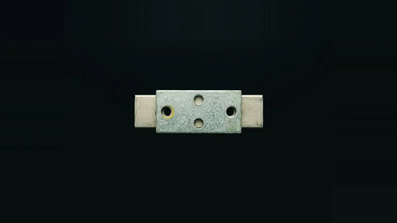 Overhead close-up of a galvanised steel splice plate on two metal bars, one bolt hole misaligned and ringed in yellow.