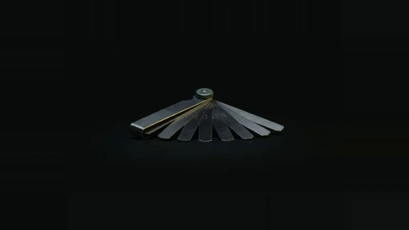 A machinist's feeler gauge fanned open into stepped steel blades, rim-lit against a black field, one edge glowing yellow.