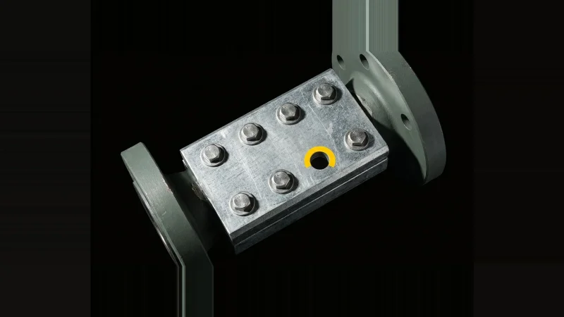 Close-up of a galvanised steel splice plate with seven bolts and one empty hole outlined in yellow on black.