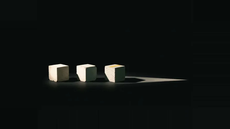 Three pale concrete test cubes in darkness; two cast short shadows while the third's shadow stretches far into black.